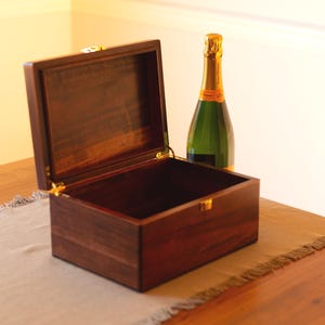 May include: A dark wood box with a hinged lid is open, revealing an empty interior. A bottle of champagne is sitting on a table next to the box.