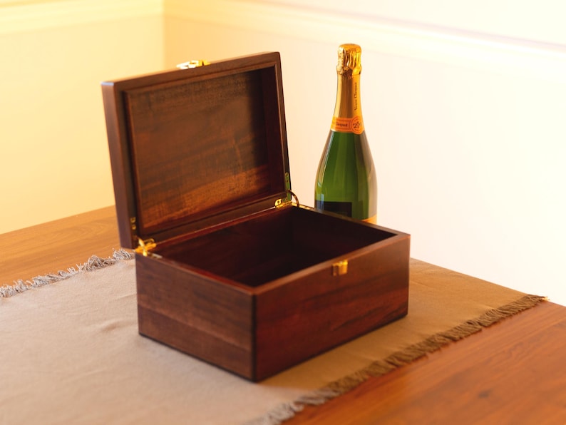 May include: A dark wood box with a hinged lid is open, revealing an empty interior. A bottle of champagne is sitting on a table next to the box.