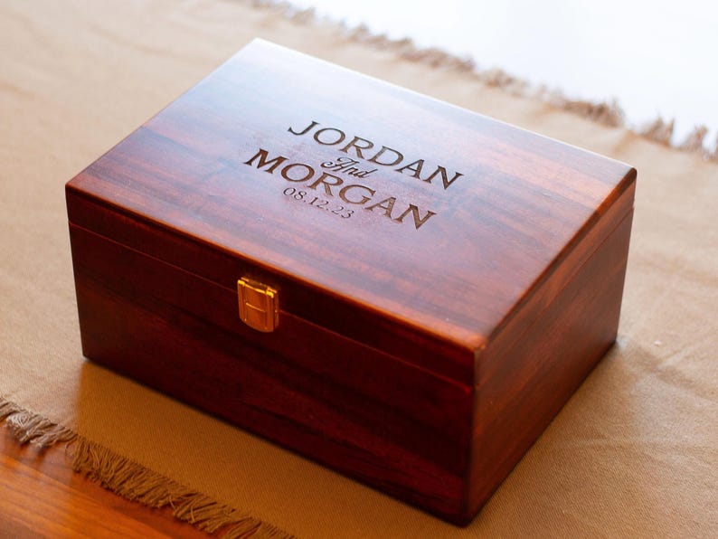 May include: A dark wood box with a brass latch. The box is engraved with the text "JORDAN and MORGAN 08.12.23".