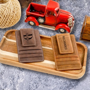 May include: Two brown leather wallets with a money clip and personalized with the names "David" and "Richard". The wallets are displayed on a wooden tray.