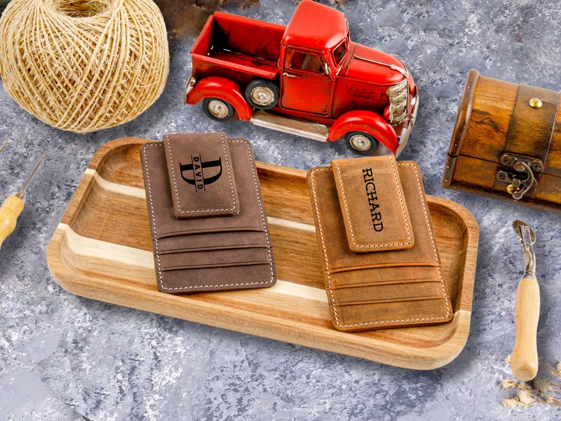May include: Two brown leather wallets with a money clip and personalized with the names "David" and "Richard". The wallets are displayed on a wooden tray.