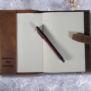 May include: A brown leather-bound journal with a pen and the inscription "Embrace the journey" on the front cover.