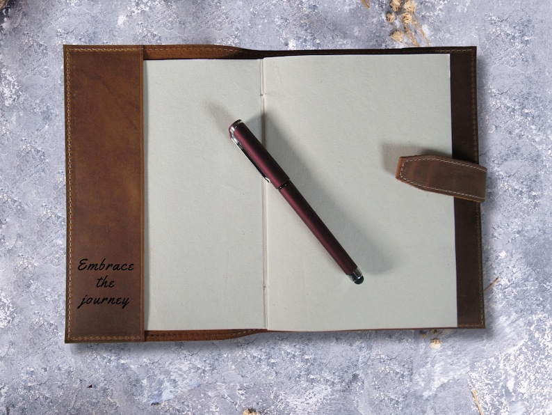 May include: A brown leather-bound journal with a pen and the inscription "Embrace the journey" on the front cover.