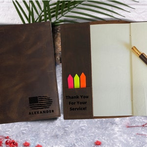 May include: A brown leather journal with a personalized inscription that reads "Thank You For Your Service!" and "ALEXANDER". The journal has a pocket on the front cover with three colorful sticky notes. The journal is open to reveal blank pages and a pen.
