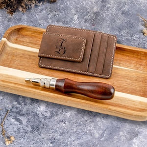 May include: A brown leather wallet with a money clip and a personalized inscription that reads "Luke". The wallet is on a wooden tray with a small tool.