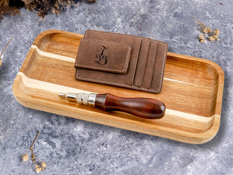 May include: A brown leather wallet with a money clip and a personalized inscription that reads "Luke". The wallet is on a wooden tray with a small tool.