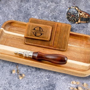 May include: A brown leather wallet with a baseball design and the name "Jacob" engraved on it. The wallet is sitting on a wooden tray with a small screwdriver.