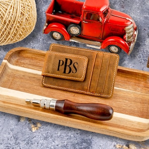 May include: A brown leather wallet with a money clip and the initials PBS embossed on the front. The wallet is sitting on a wooden tray with a red toy truck and a ball of twine in the background.