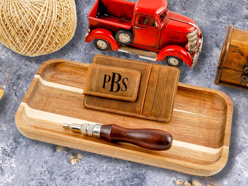 May include: A brown leather wallet with a money clip and the initials PBS embossed on the front. The wallet is sitting on a wooden tray with a red toy truck and a ball of twine in the background.