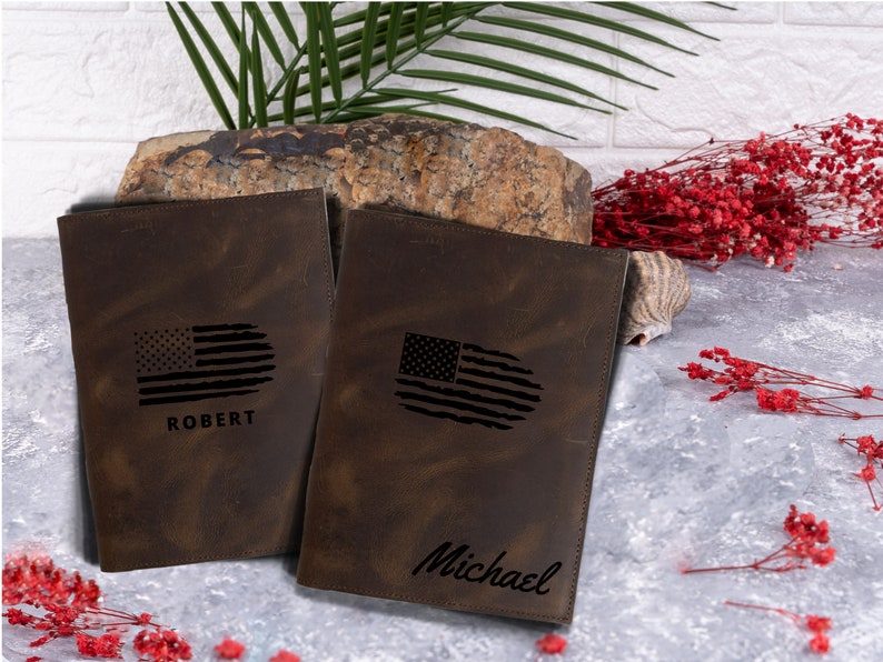 May include: Two brown leather journals with distressed finishes. Each journal features a black and white American flag design and a personalized name. The names are "Robert" and "Michael".