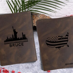 May include: Two brown leather journals with personalized engravings. The left journal features a black silhouette of the New York City skyline with the Statue of Liberty and the name "BRUCE". The right journal features a black silhouette of the United States with an American flag design and the name "RICHARD".