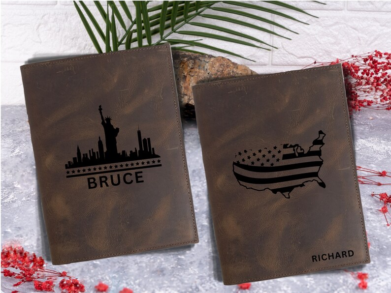 May include: Two brown leather journals with personalized engravings. The left journal features a black silhouette of the New York City skyline with the Statue of Liberty and the name "BRUCE". The right journal features a black silhouette of the United States with an American flag design and the name "RICHARD".