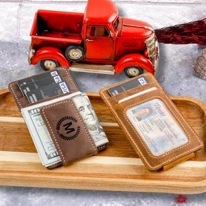 May include: Two brown leather wallets with a money clip and a clear ID window. One wallet has a monogrammed "M" with a laurel wreath design. The wallets are on a wooden tray with a red toy truck in the background.