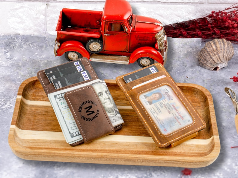 May include: Two brown leather wallets with a money clip and a clear ID window. One wallet has a monogrammed "M" with a laurel wreath design. The wallets are on a wooden tray with a red toy truck in the background.