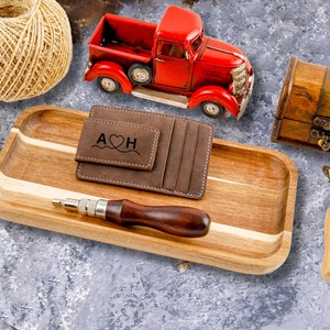 May include: A brown leather wallet with a money clip and the initials "A&hearts;H" engraved on the front. The wallet is on a wooden tray with a red toy truck, a ball of twine, and a small wooden chest.