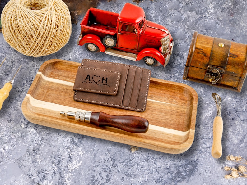 May include: A brown leather wallet with a money clip and the initials "A&hearts;H" engraved on the front. The wallet is on a wooden tray with a red toy truck, a ball of twine, and a small wooden chest.