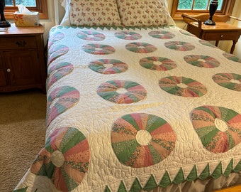 May include: A white bed with a white and green quilt with a floral pattern. The quilt has a scalloped edge. The bed has a white headboard and a white footboard. There are two white pillows with floral patterns on the bed. The bed is in a bedroom with a window in the background.