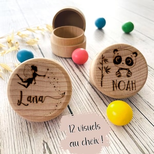 Op de afbeelding: Houten bewaardoosjes met gegraveerde ontwerpen. Eén doosje heeft een fee met de naam "Lana", een ander een panda met de naam "Noah". Het open doosje is zichtbaar, samen met kleurrijke bollen. De tekst "12 visuels au choix !" is ook aanwezig.