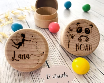 May include: Wooden keepsake boxes with engraved designs. One box features a fairy with the name "Lana", another a panda with the name "Noah". The open box is visible, along with colorful spheres. The text "12 visuals au choix !" is also present.