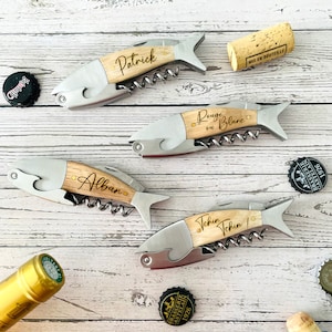 Könnte beinhalten: Vier fischförmige Korkenzieher mit Holzgriffen und Edelstahlkörpern werden gezeigt. Jeder Korkenzieher hat einen eingravierten Namen: "Patrick", "Alban", "Tchin Tchin" und "Rouge ou Blanc". Flaschendeckel und ein Korken sind verstreut.