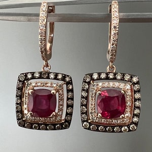 May include: A pair of rose gold earrings with a square-shaped design. Each earring features a red gemstone surrounded by a halo of smaller diamonds and brown diamonds.