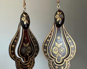 May include: A pair of ornate, drop earrings with a black and gold floral design. The earrings feature an oval top with a floral pattern and a teardrop-shaped bottom with intricate gold detailing. The earrings are suspended from gold-colored ear wires.