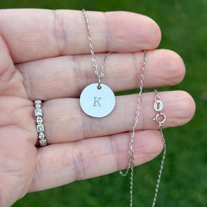 May include: A silver necklace with a delicate chain and a circular pendant. The pendant is engraved with the letter "K". A ring with square-cut stones is also visible.