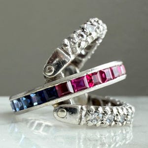May include: Three silver-toned hoop earrings. One features a row of blue gemstones, another a row of red gemstones, and the third is adorned with small, clear gemstones. The earrings are interlocked, creating a layered effect.