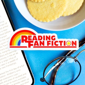 May include: A close-up of a sticker with a rainbow and the text "Reading Fan Fiction" on a blue background. There is a smartphone with a book open on the screen, a plate of cookies, and a pair of glasses.