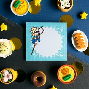 May include: A blue and white notepad with a cartoon illustration of a girl with blonde hair holding a staff. The notepad is surrounded by various small, colorful objects, including a donut, a bowl of noodles, and a bowl of dumplings.