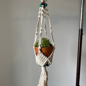 May include: A white macrame plant hanger with a wooden ring at the top and a tassel at the bottom. The hanger is decorated with brown and teal beads. A small terracotta pot with a succulent plant is hanging in the macrame.
