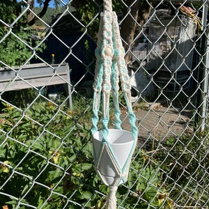 May include: A macrame plant hanger with a white and light blue design. The hanger is made of cotton cord and has a white pot attached to it. The hanger is hanging from a chain link fence.