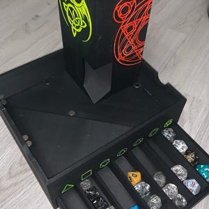 May include: Black dice tower and storage box with a drawer. The tower features a neon green dragon design and a red circular design. The box contains various polyhedral dice in different colours and styles. A gaming accessory.