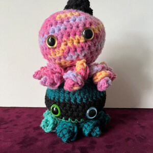 May include: Two crocheted octopus plushies stacked on top of each other. The top octopus is pink, yellow, and purple with a black hat. The bottom octopus is black, teal, and green.