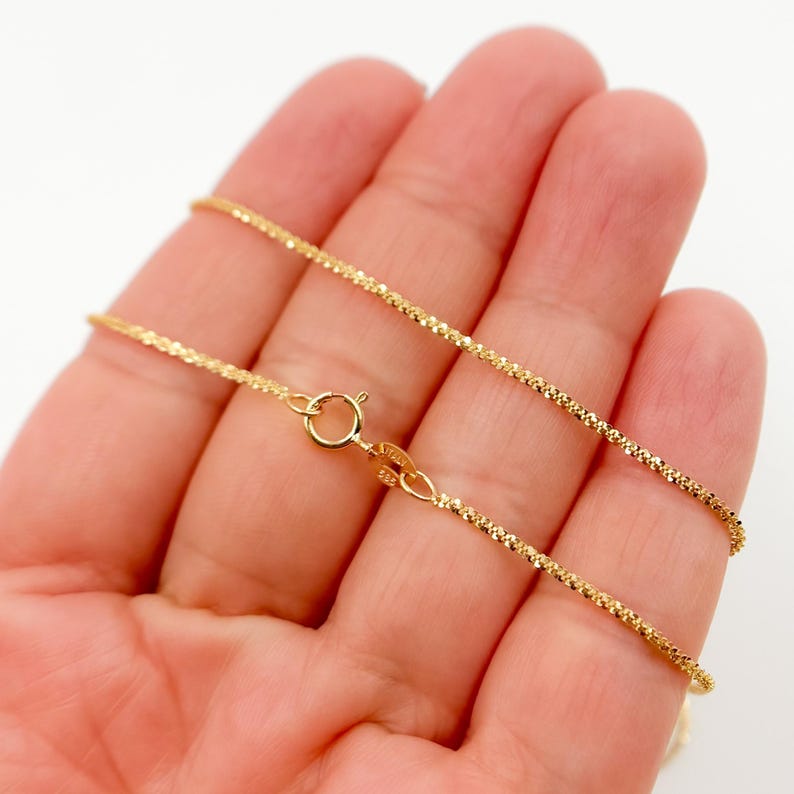 May include: A gold chain necklace with a delicate, twisted design. The chain is shown on a hand, and the clasp is visible.