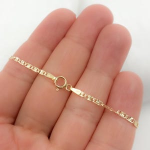 May include: A delicate gold-colored chain necklace with a lobster clasp. The chain has a repeating pattern of small, diamond-shaped links. The necklace is shown against a light background.