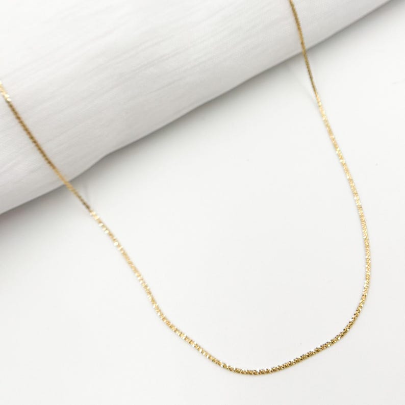 May include: A delicate gold chain necklace with a fine, textured design. The chain is thin and has a subtle shimmer.