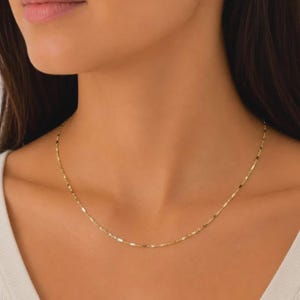 May include: A delicate gold chain necklace featuring a series of small, flat, rectangular links connected by thin circular rings. The necklace rests against the neck, showcasing a polished, reflective metallic finish with a consistent, repeating pattern.
