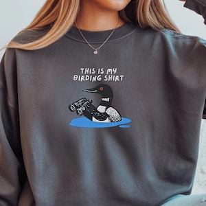 May include: Dark gray sweatshirt with the text "THIS IS MY BIRDING SHIRT" above an embroidered cartoon of a loon holding binoculars in a blue puddle. The loon is black, white, and gray.