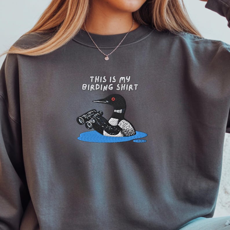 May include: Dark gray sweatshirt with the text "THIS IS MY BIRDING SHIRT" above an embroidered cartoon of a loon holding binoculars in a blue puddle. The loon is black, white, and gray.