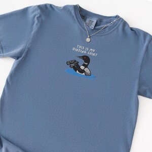 May include: A blue short-sleeved t-shirt with the text "THIS IS MY BIRDING SHIRT" above a cartoon of a loon holding binoculars. The loon is black, white, and red, and is in blue water. A silver necklace is on the shirt.