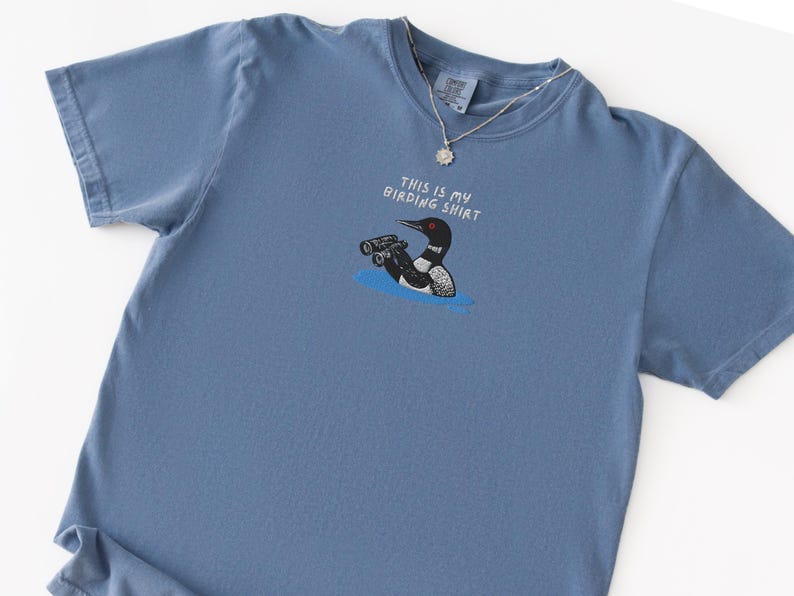 May include: A blue short-sleeved t-shirt with the text "THIS IS MY BIRDING SHIRT" above a cartoon of a loon holding binoculars. The loon is black, white, and red, and is in blue water. A silver necklace is on the shirt.