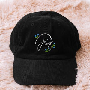 May include: Black corduroy baseball cap with a white embroidered manatee design and blue floral accents. The cap has a curved peak and an adjustable strap. A fashionable accessory for everyday wear.
