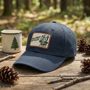 May include: A navy blue baseball cap with a patch featuring an owl illustration and the text "GIVE A HOOT! DON'T POLLUTE". The cap is on a wooden surface with a mug and pinecones in the background.