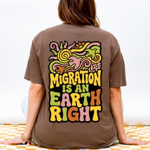 Peut inclure: Un t-shirt marron avec un motif coloré au dos. Le motif représente un soleil, des fleurs et des formes abstraites en jaune, vert, rose et orange, avec le texte "MIGRATION IS AN EARTH RIGHT".