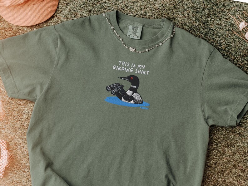 May include: Olive green t-shirt with the text "THIS IS MY BIRDING SHIRT" and a graphic of a loon holding binoculars. The shirt has a crew neck and short sleeves. A silver necklace with small beads and butterfly charms is around the collar.