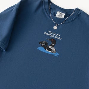 May include: A navy blue t-shirt with the text "THIS IS MY BIRDING SHIRT" and a cartoon illustration of a loon with binoculars. A silver necklace with a pendant is draped on the shirt.