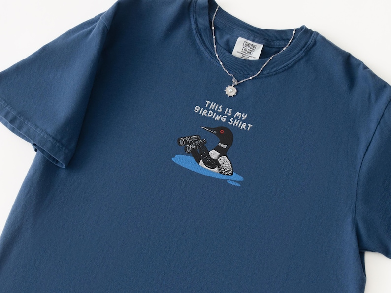 May include: A navy blue t-shirt with the text "THIS IS MY BIRDING SHIRT" and a cartoon illustration of a loon with binoculars. A silver necklace with a pendant is draped on the shirt.