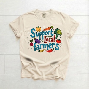 May include: A cream-coloured short-sleeve t-shirt featuring the blue and red text "Support Local farmers" surrounded by colourful illustrations of a yellow bell pepper, orange carrot, green broccoli, purple aubergine, red radish, tomato, and chilli pepper.