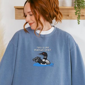 May include: A blue sweatshirt with the text "THIS IS MY BIRDING SHIRT" above an illustration of a bird with binoculars. The bird is black and white with a red eye, sitting in a blue puddle.